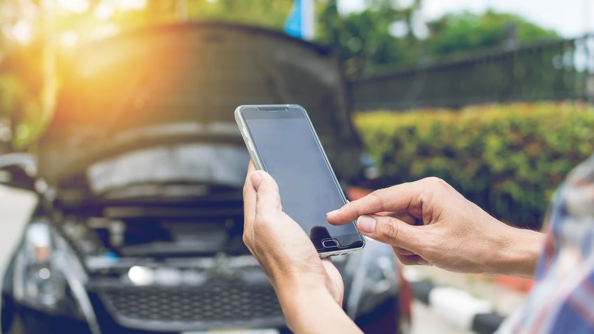 A driver using a mobile phone to call for help in front of a car with its hood up, demonstrating the need for vehicle service protection and roadside assistance.