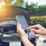 A driver using a mobile phone to call for help in front of a car with its hood up, demonstrating the need for vehicle service protection and roadside assistance.