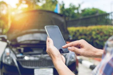 A driver using a mobile phone to call for help in front of a car with its hood up, demonstrating the need for vehicle service protection and roadside assistance.